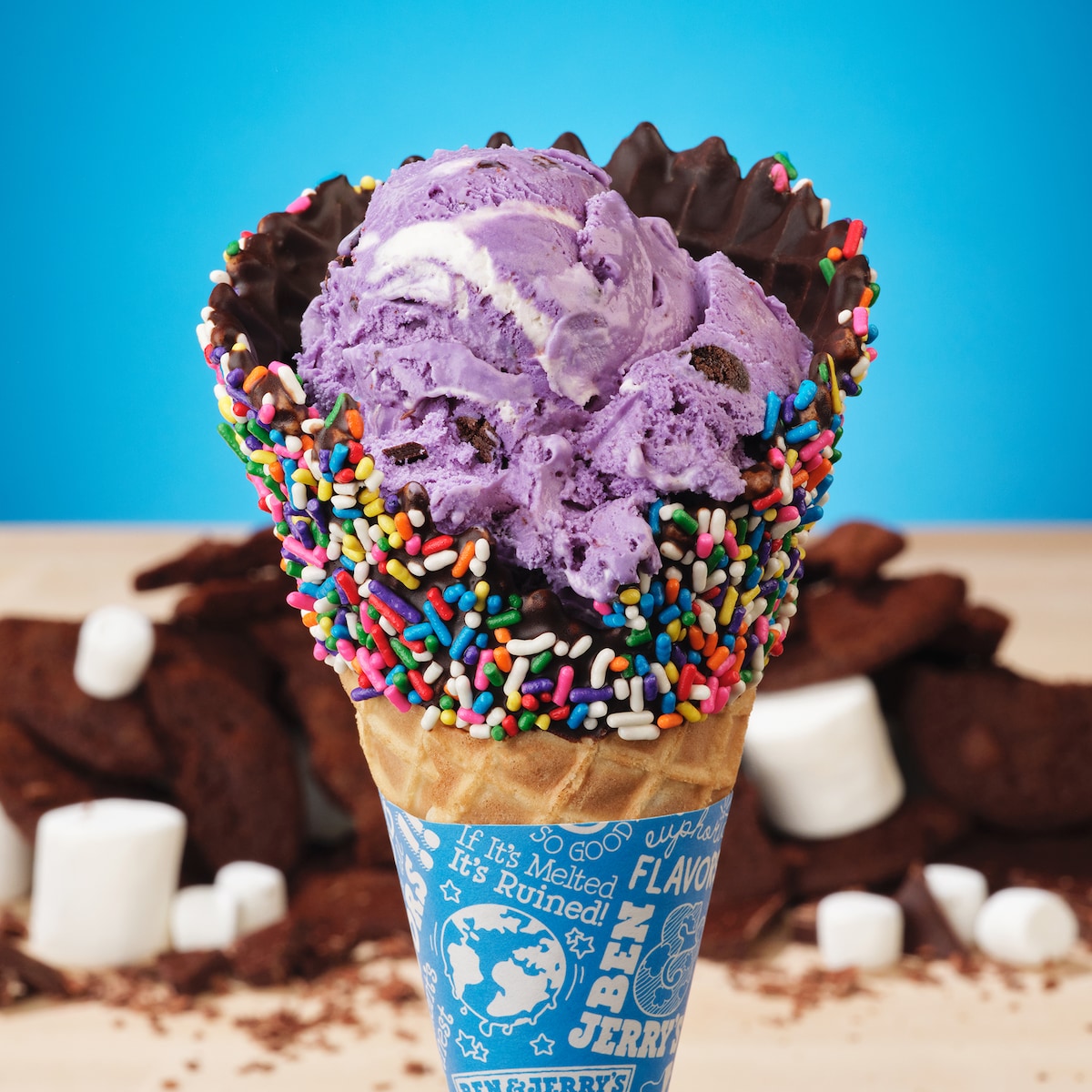 Chocolate-dipped waffle cone with Ben & Jerry's UltraViolet ice cream inside. In the background, brownies and marshmallows.