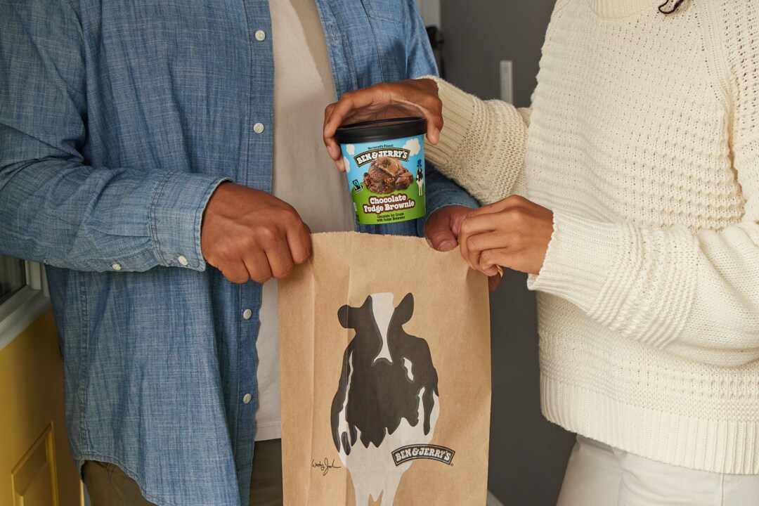 Two people holding a Ben & Jerry's delivery bag, pulling a pint of Ben & Jerry's Chocolate Fudge Brownie ice cream out of the bag.