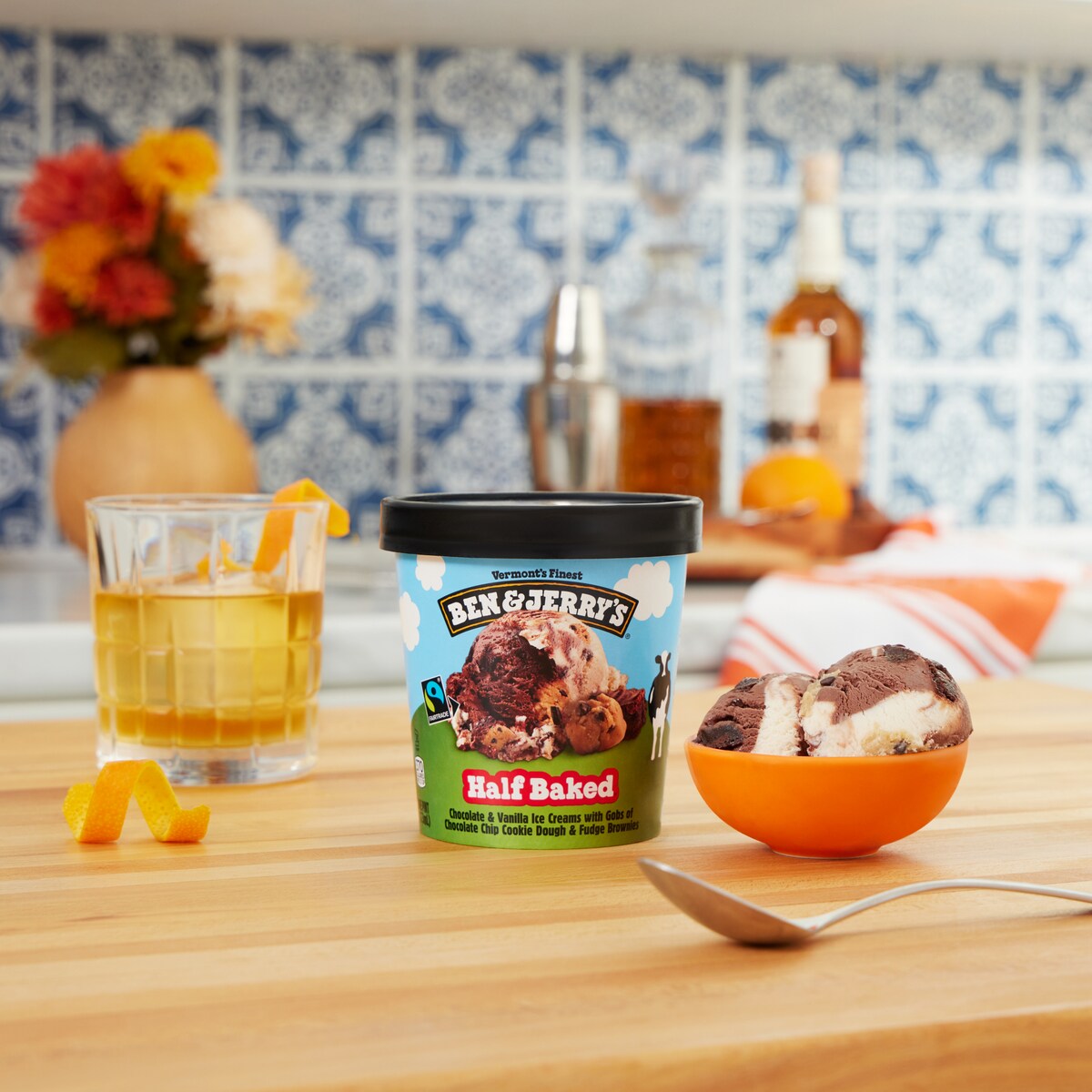 Pint of Ben & Jerry's Half Baked ice cream next to a whiskey cocktail with an orange garnish in a kitchen setting.