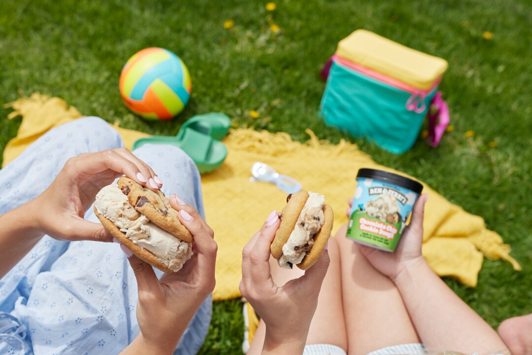 Hands holding ice cream sandwiches in an outdoor summer setting. Pint of Ben & Jerry's Chocolate Chip Cookie Dough ice cream nearby.