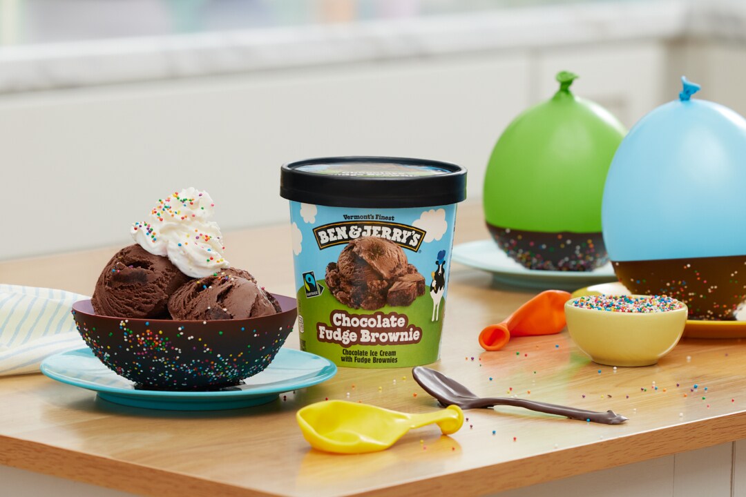 Pint of Ben & Jerry's Chocolate Fudge Brownie ice cream next to a chocolate sundae made in a chocolate bowl. Balloons nearby.